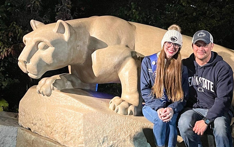 Smiling man and woman in Penn State gear sitting in front of the Nittany Lion Shrine.