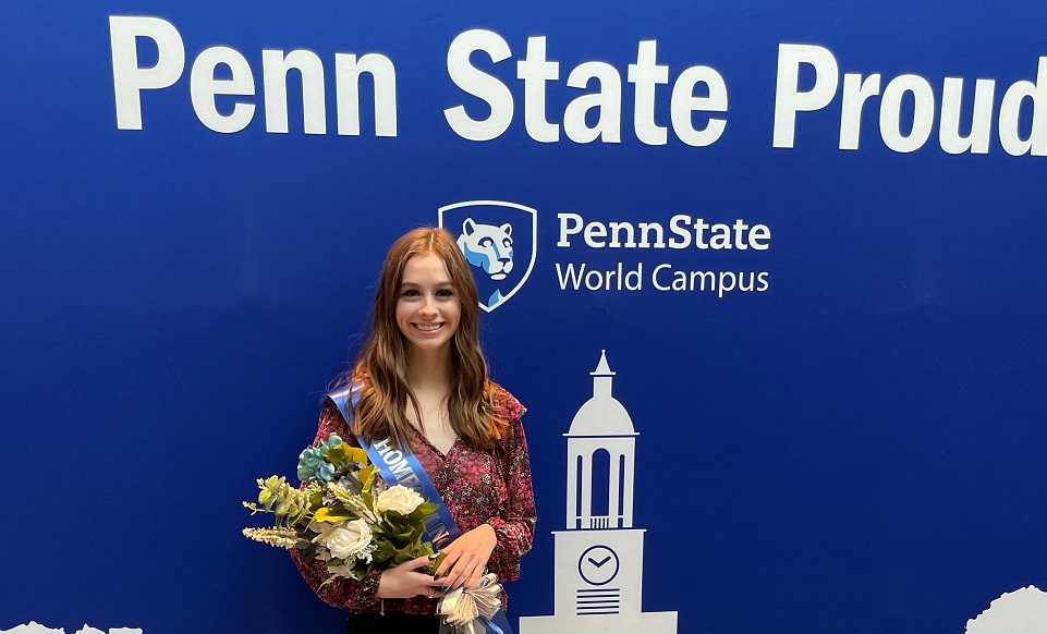 Smiling woman with long brown hair standing in front of banner with Penn State World Campus logo that says, 'Penn State Proud"