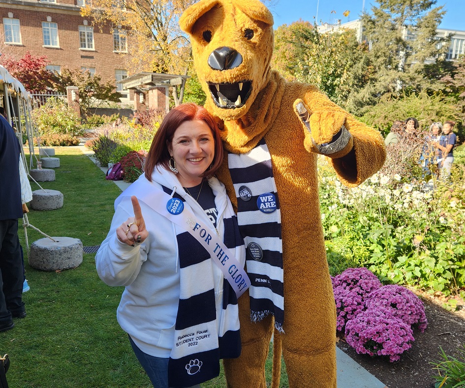 Smiling woman wearing Penn State gear and a "For the Glory" sash standing next to the Nittany Lion Mascot