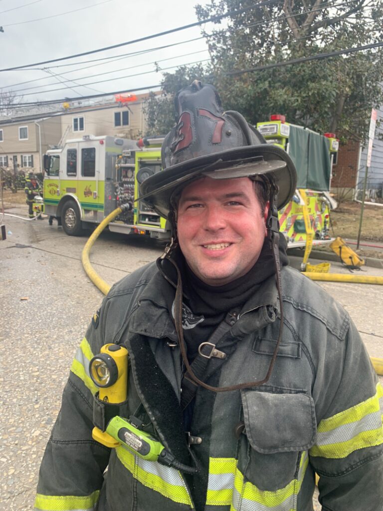 Smiling man wearing firefighter gear with fire truck in background