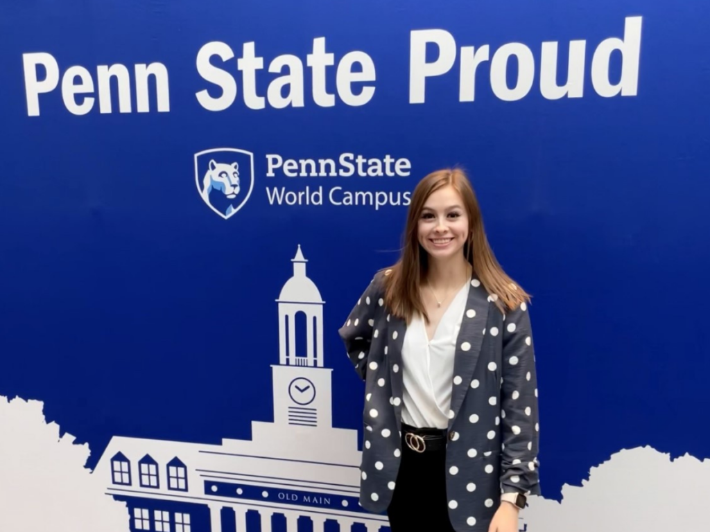 Smiling woman with long dark hair standing in front of banner with illustration of Old Main and the words "Penn State Proud" above the World Campus logo