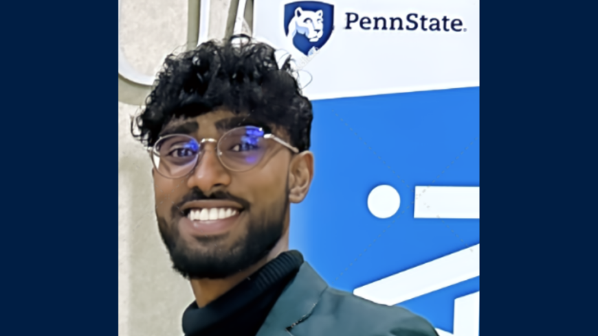 Smiling man with dark hair and glasses standing in front of banner with Penn State logo