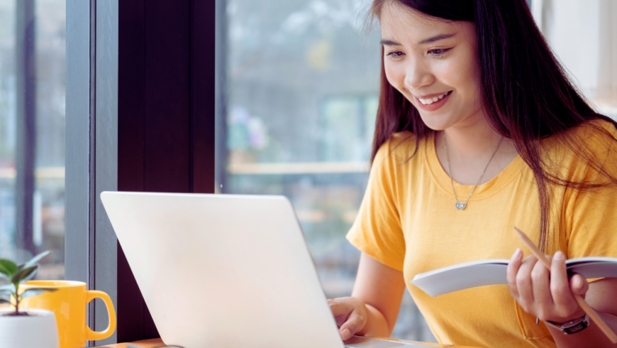 Smiling young woman with long dark hair holding a notebook while looking at a laptop screen