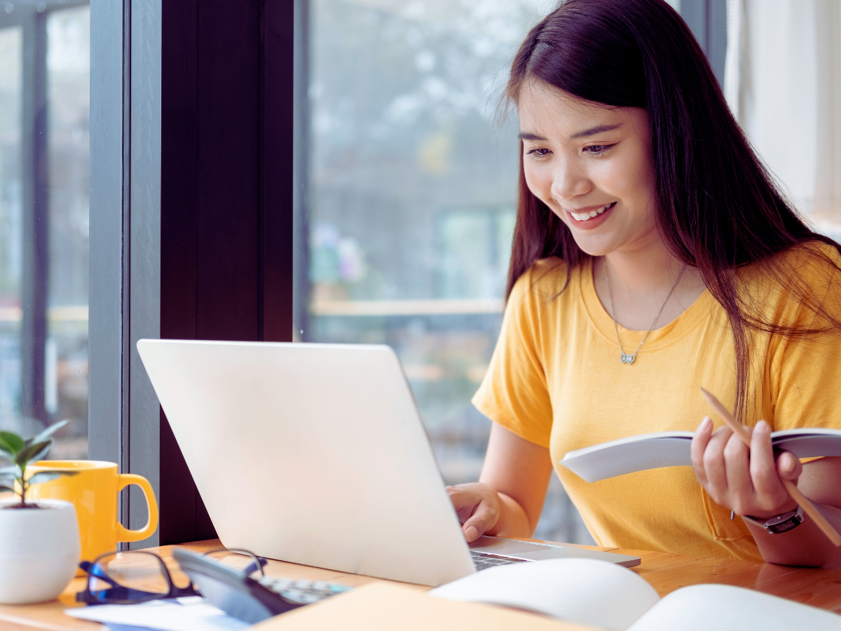 Smiling young woman with long dark hair holding a notebook while looking at a laptop screen