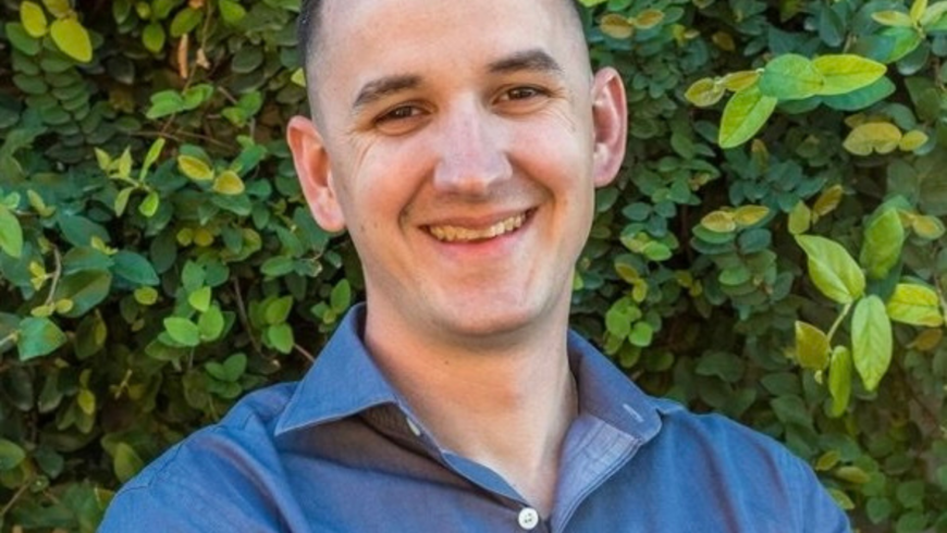 Smiling man with short dark hair wearing blue shirt and standing in front of trees in the background