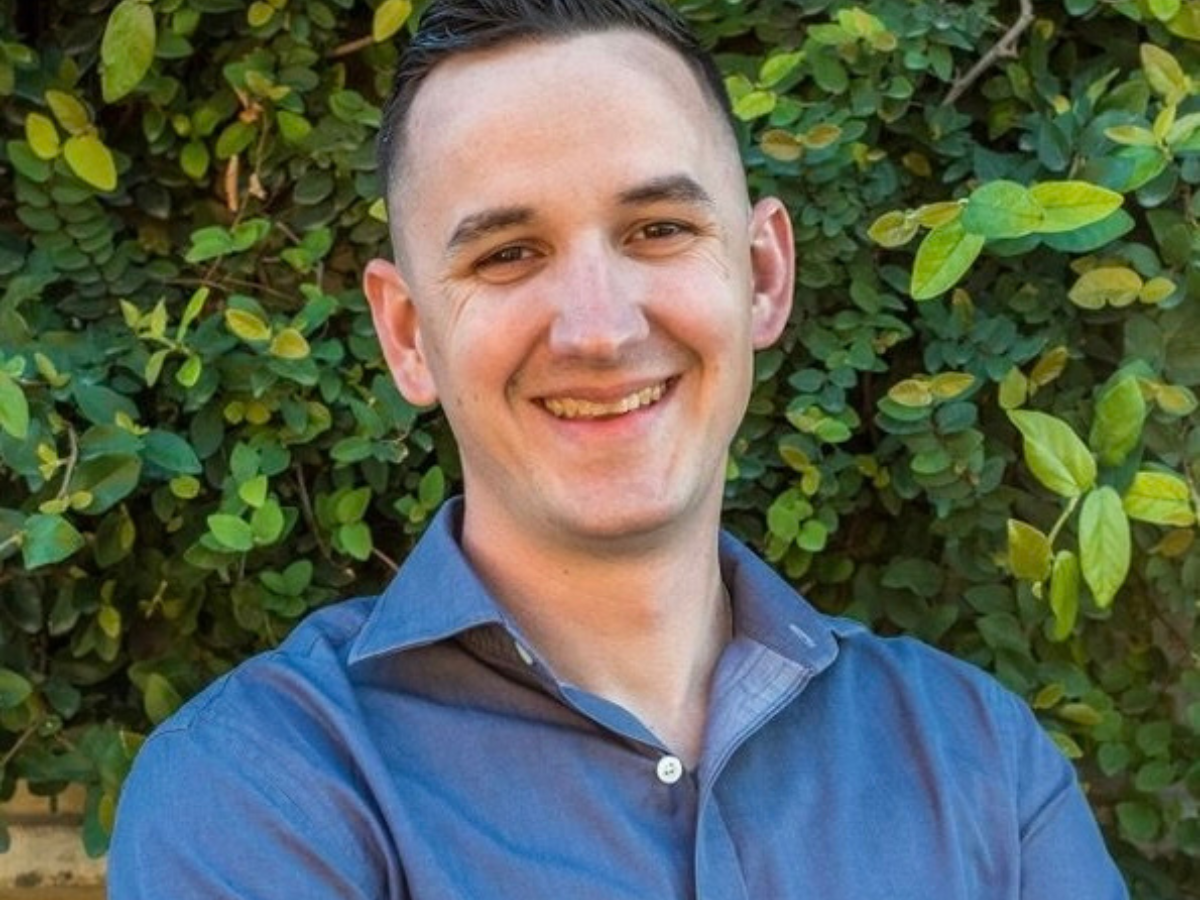 Smiling man with short dark hair wearing blue shirt and standing in front of trees in the background