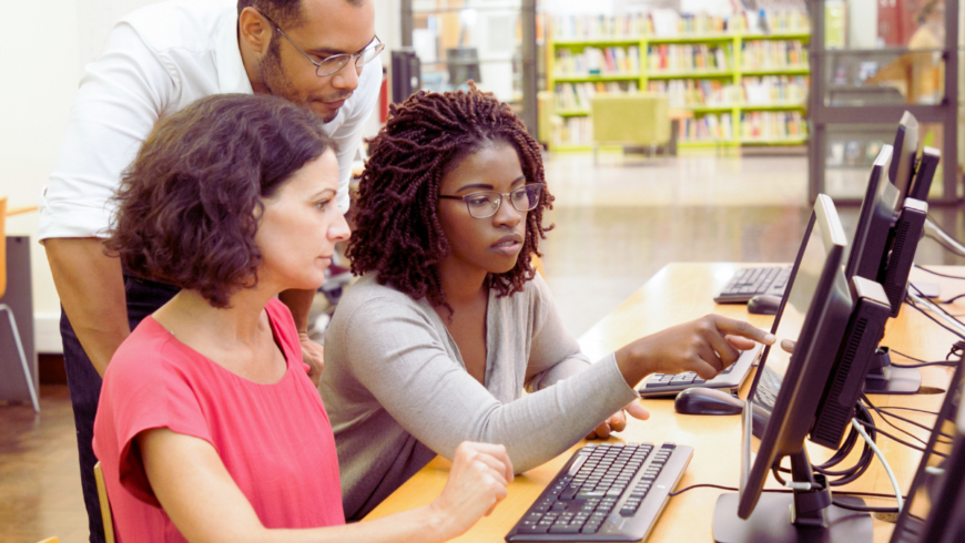 Three people – two women sitting at a table and a man leaning over their shoulders behind them – looking at a computer screen in what appears to be a computer lab at a library