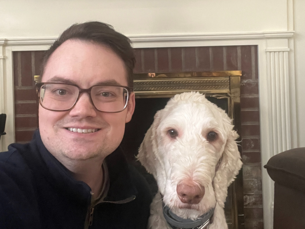 Smiling man with short brown hair wearing glasses and sitting next to a white dog