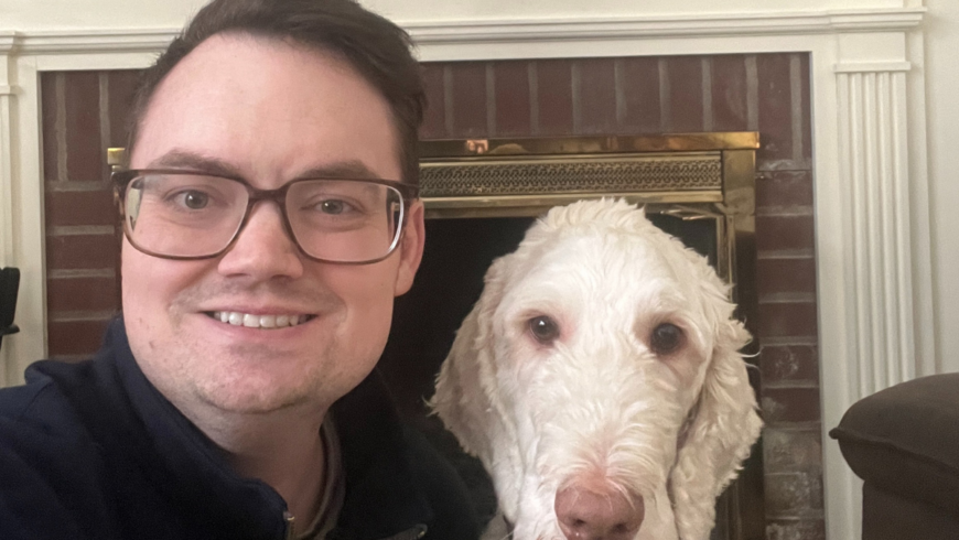 Smiling man with short brown hair wearing glasses and sitting next to a white dog